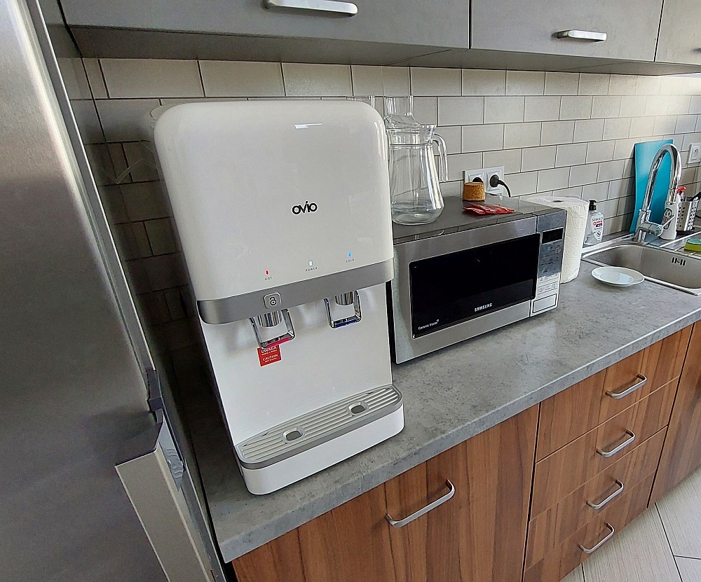 Dystrybutor wody marki Ovio na blacie kuchennym obok mikrofalówki, z widocznymi przyciskami do wody zimnej i gorącej oraz ostrzeżeniem o gorącej wodzie.
