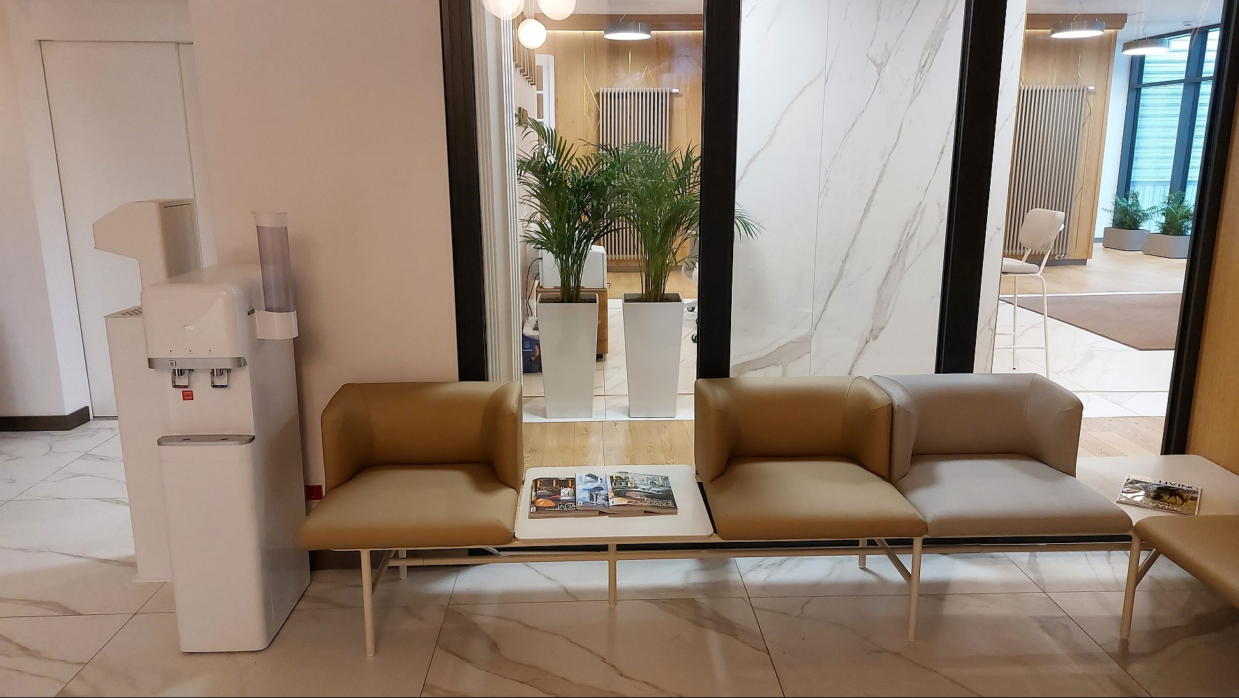 Wnętrze biura z dystrybutorem wody, roślinami w donicach i miejscami do siedzenia z gazetami.