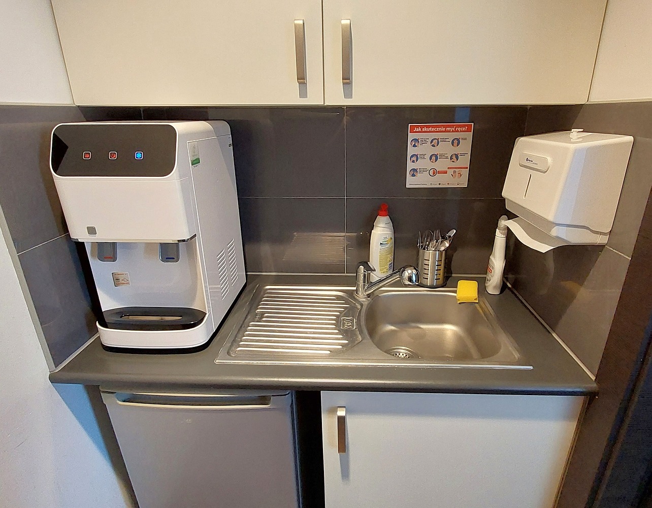 Kompaktowy dystrybutor wody z trzema przyciskami i zintegrowanym zlewem, umieszczony na szafce pod zlewem w biurze, obok dozownika ręczników papierowych i instrukcji mycia rąk.