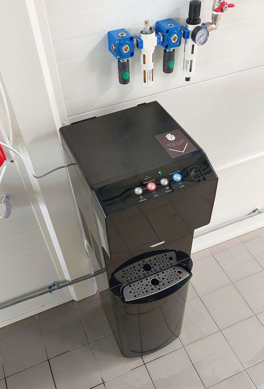 Czarny dystrybutor wody z przyciskami do wyboru temperatury, zamontowany w pomieszczeniu z szarą podłogą i białymi ścianami, nad nim filtry przemysłowe.