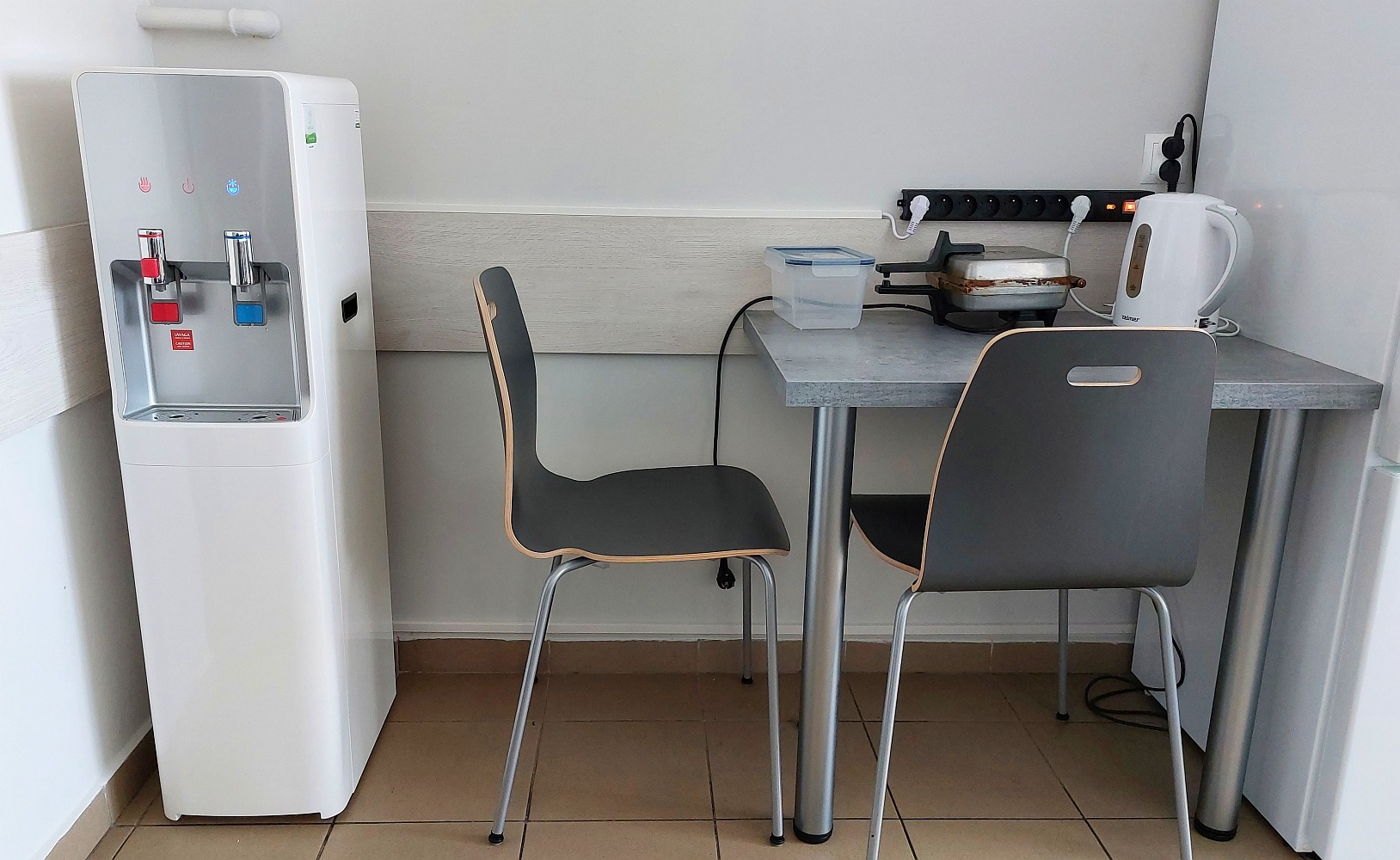 Biały dystrybutor wody z kranikami na ciepłą i zimną wodę, stojący obok szarego stołu z czajnikiem elektrycznym i opiekaczem do kanapek w pomieszczeniu biurowym.