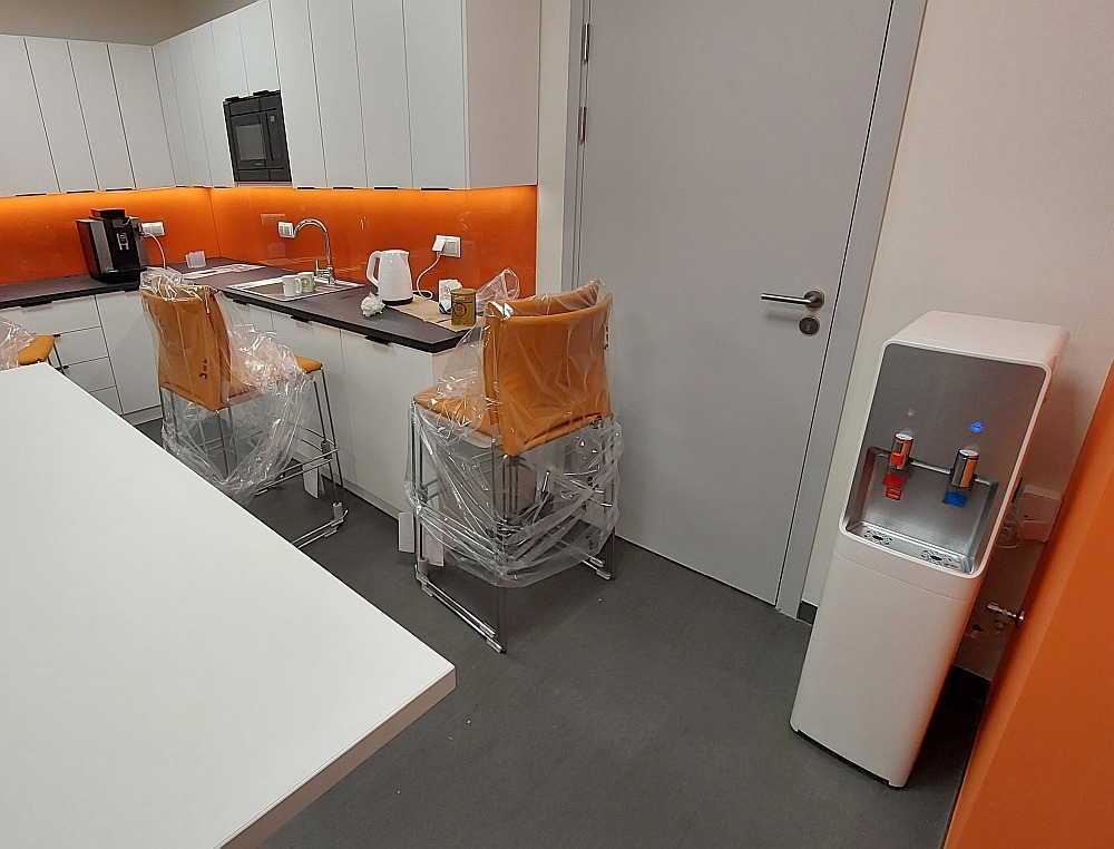 Nowoczesna kuchnia biurowa z białymi szafkami, pomarańczowym panelem, krzesłami barowymi w folii ochronnej i dystrybutorem wody.