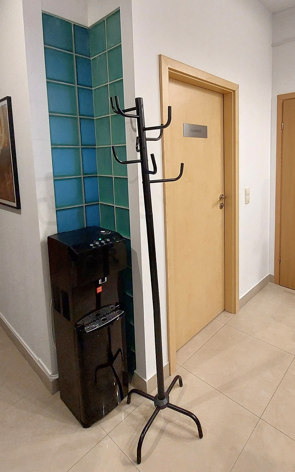 Czarny dystrybutor wody z widocznymi przyciskami i wskaźnikami LED stoi obok ściany z niebiesko-zielonych szklanych płytek, a obok niego wieszak na ubrania.