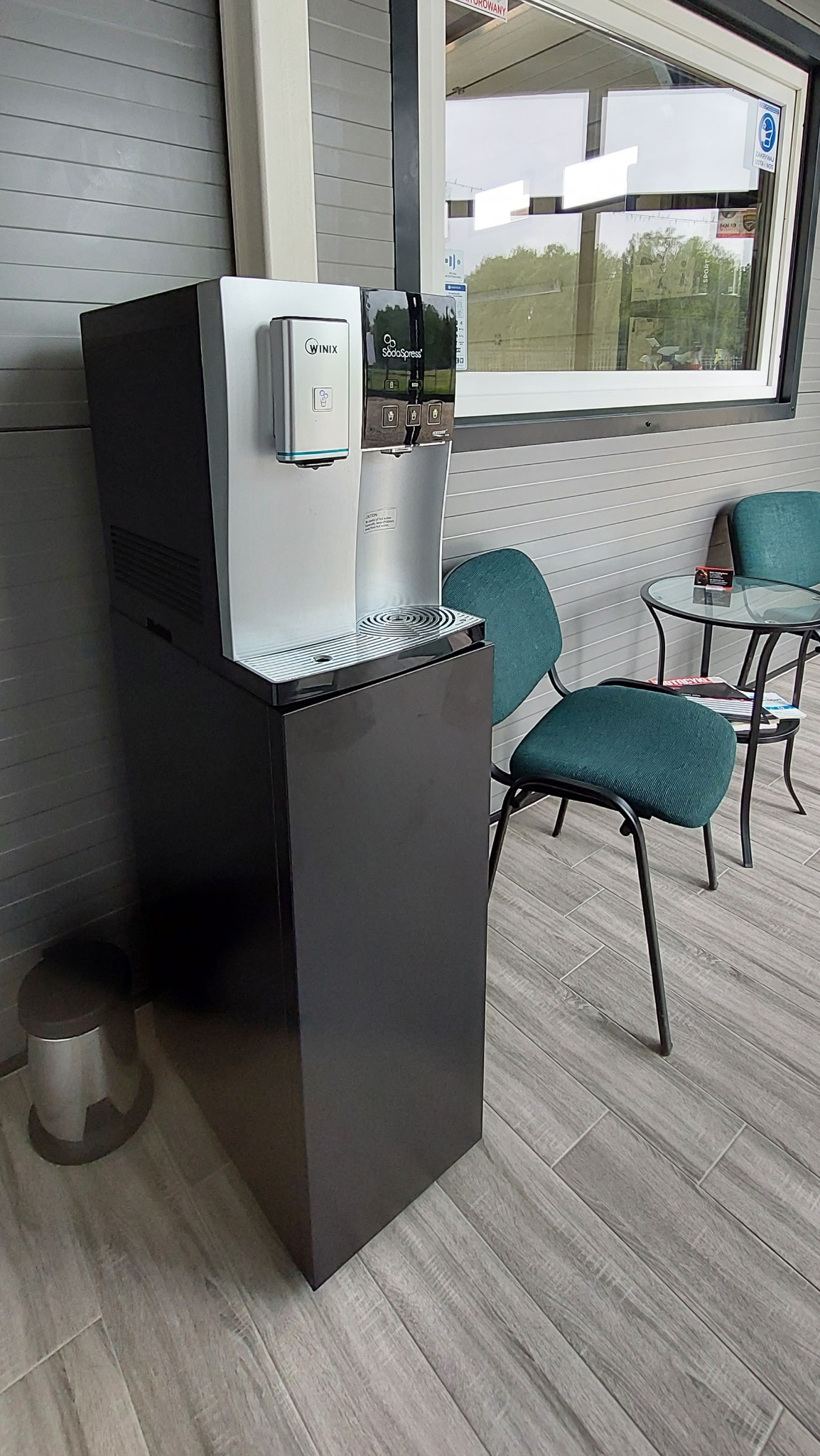 Stojący dystrybutor wody Winix SodaSpress w pomieszczeniu biurowym z krzesłami i stolikiem, widok z boku.