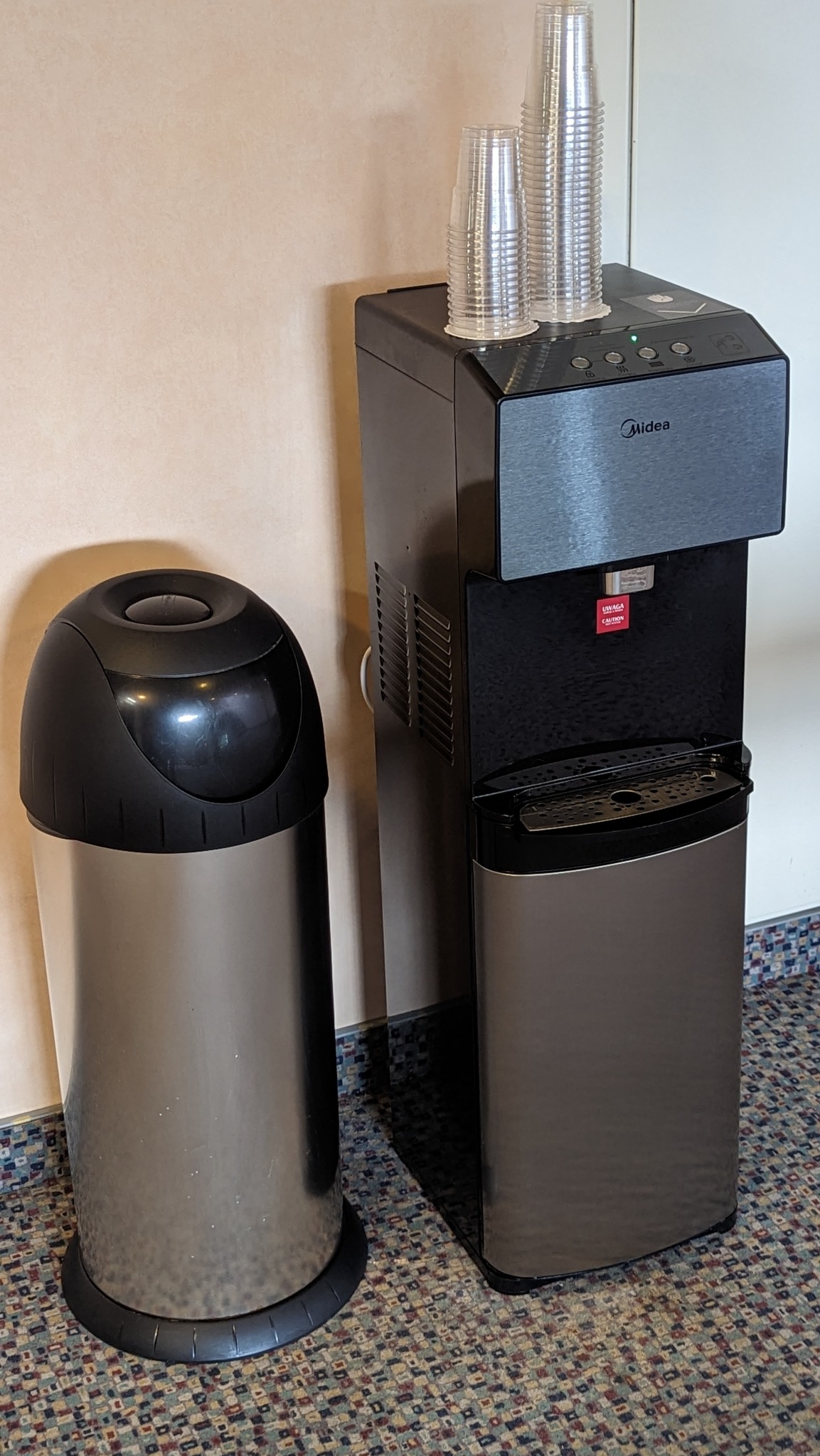 Dystrybutor wody Midea w biurowym otoczeniu, obok kosza na śmieci i z zapasem plastikowych kubków na górze.