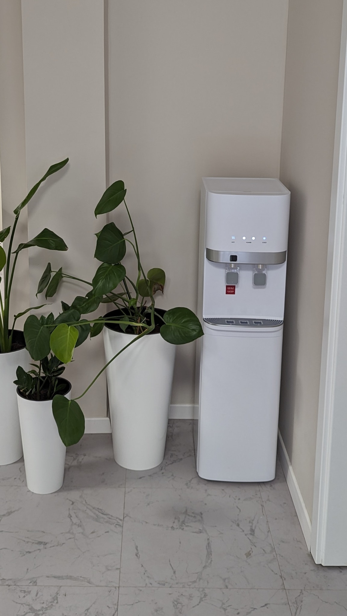 Nowoczesny, biały dystrybutor wody z butelką, ustawiony obok dwóch białych doniczek z zielonymi roślinami na tle jasnoszarej ściany i marmurowej podłogi.