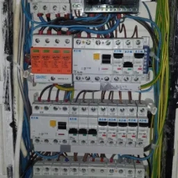 Rozdzielnica elektryczna z bezpiecznikami EATON i SIMTEC, okablowaniem w kolorach niebieskim, zielono-żółtym i brązowym, zasilana przewodami. Widoczne zabezpieczenia przeciwprzepięciowe.