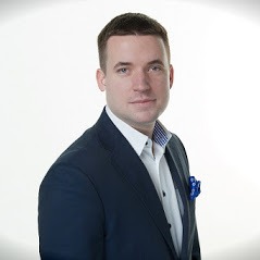 Portret mężczyzny w ciemnym garniturze i błękitnej poszetce, na jasnym tle, z widocznym kołnierzykiem koszuli w paski.