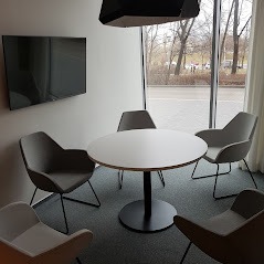 Nowoczesne wnętrze biurowe z okrągłym stołem konferencyjnym i krzesłami w odcieniach szarości, oświetlone naturalnym światłem z dużego okna.