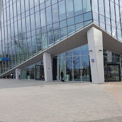 Nowoczesny biurowiec z przeszkloną fasadą, widok z zewnątrz, jasny chodnik, obok wejścia widoczny szyld z nazwą firmy.