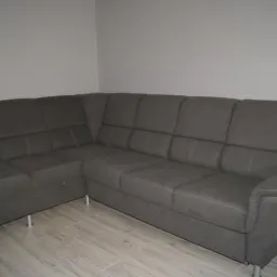 Szary narożnik z funkcją spania i pojemnikiem na pościel, widoczne metalowe nóżki i przeszycia na siedzisku, ustawiony na panelach podłogowych w odcieniach szarości.