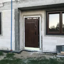 Brązowe drzwi wejściowe w stanie surowym, osadzone w ścianie z betonu komórkowego, widoczne ślady pianki montażowej i obróbki tynkarskiej.