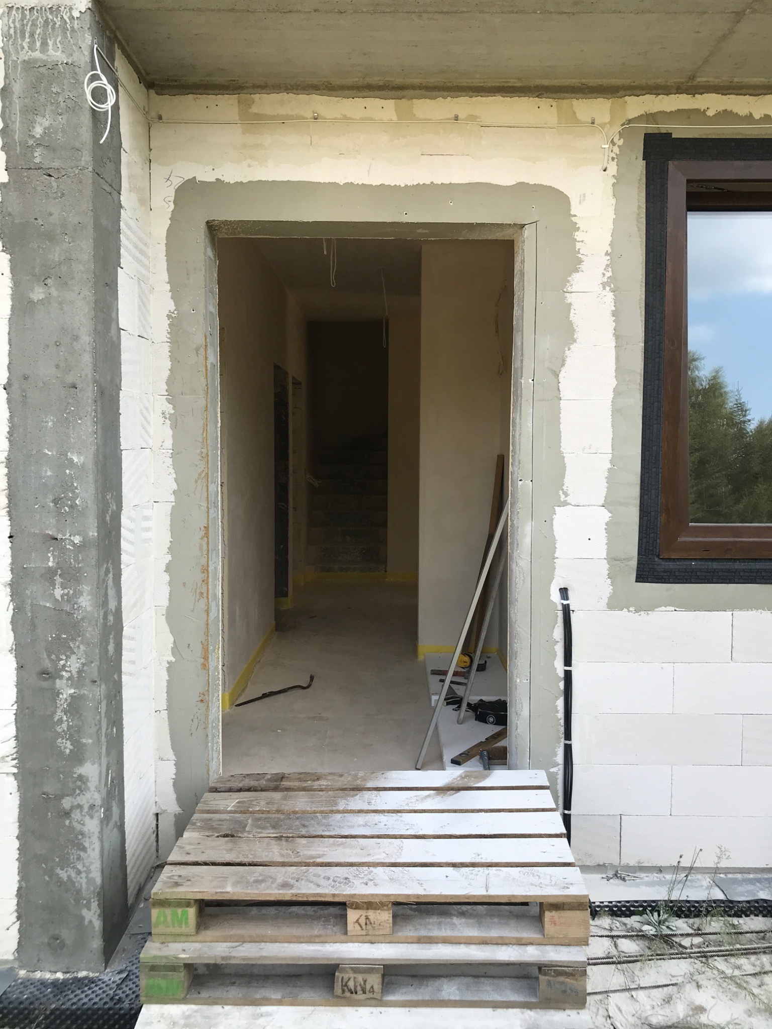 Surowy stan wykończenia otworu drzwiowego w Warszawie, widoczny fragment ściany z betonu komórkowego, częściowo otynkowana, paleta drewniana jako tymczasowe wejście, w tle schody.