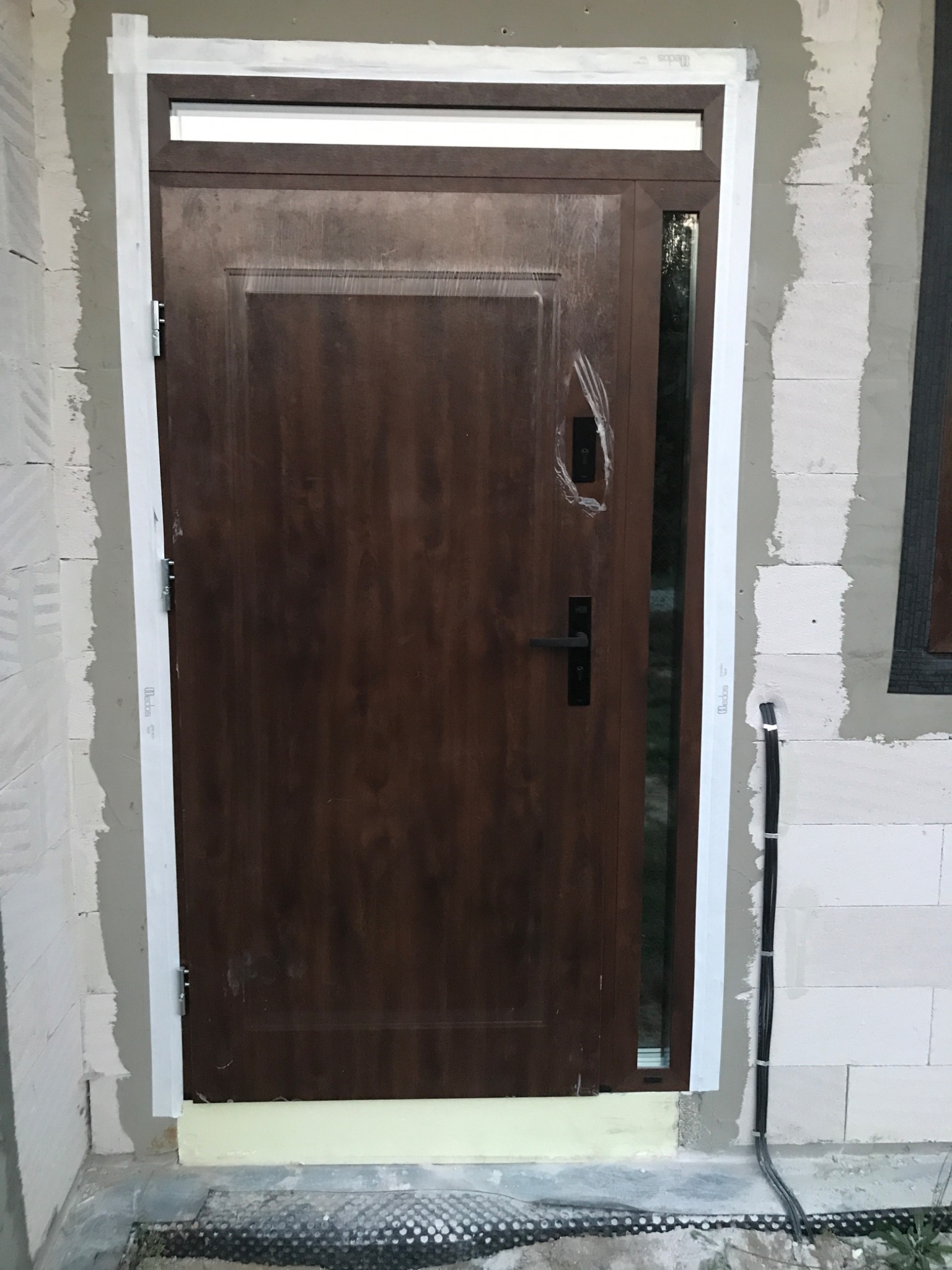 Brązowe drzwi wejściowe z bocznym przeszkleniem i górnym naświetlem, osadzone w surowym otworze, zabezpieczone folią, z widocznymi przewodami elektrycznymi po prawej stronie.