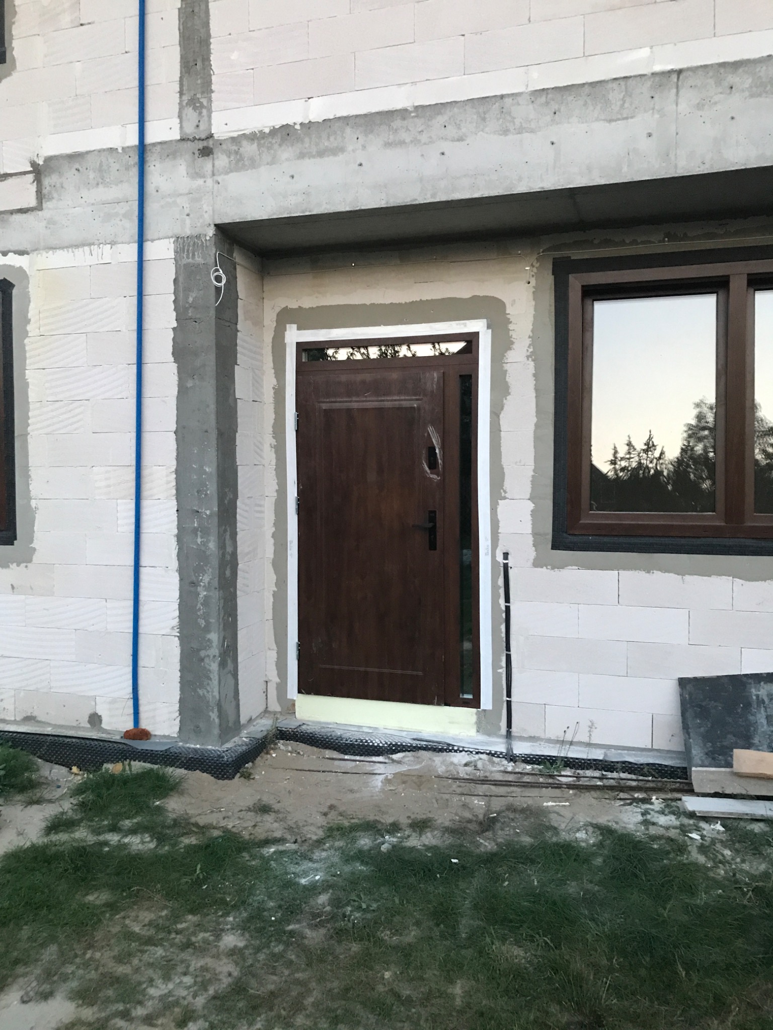 Brązowe drzwi wejściowe w stanie surowym, osadzone w ścianie z betonu komórkowego, widoczne ślady pianki montażowej i obróbki tynkarskiej.