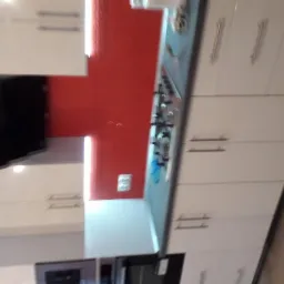 Nowoczesna kuchnia z białymi szafkami, czerwonym panelem ściennym nad kuchenką gazową i czarnym okapem.