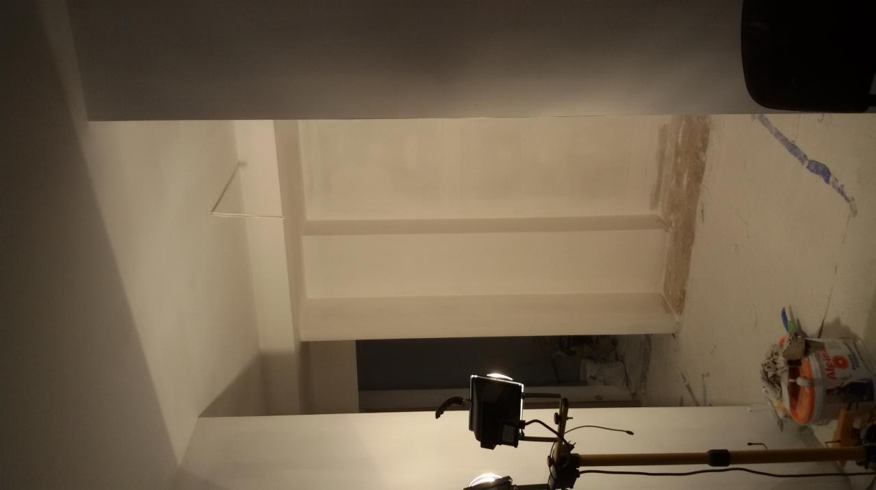 Wnętrze po malowaniu: gładkie, białe ściany i sufit, oświetlone lampą budowlaną. Widoczne narzędzia malarskie i wiadro z farbą.