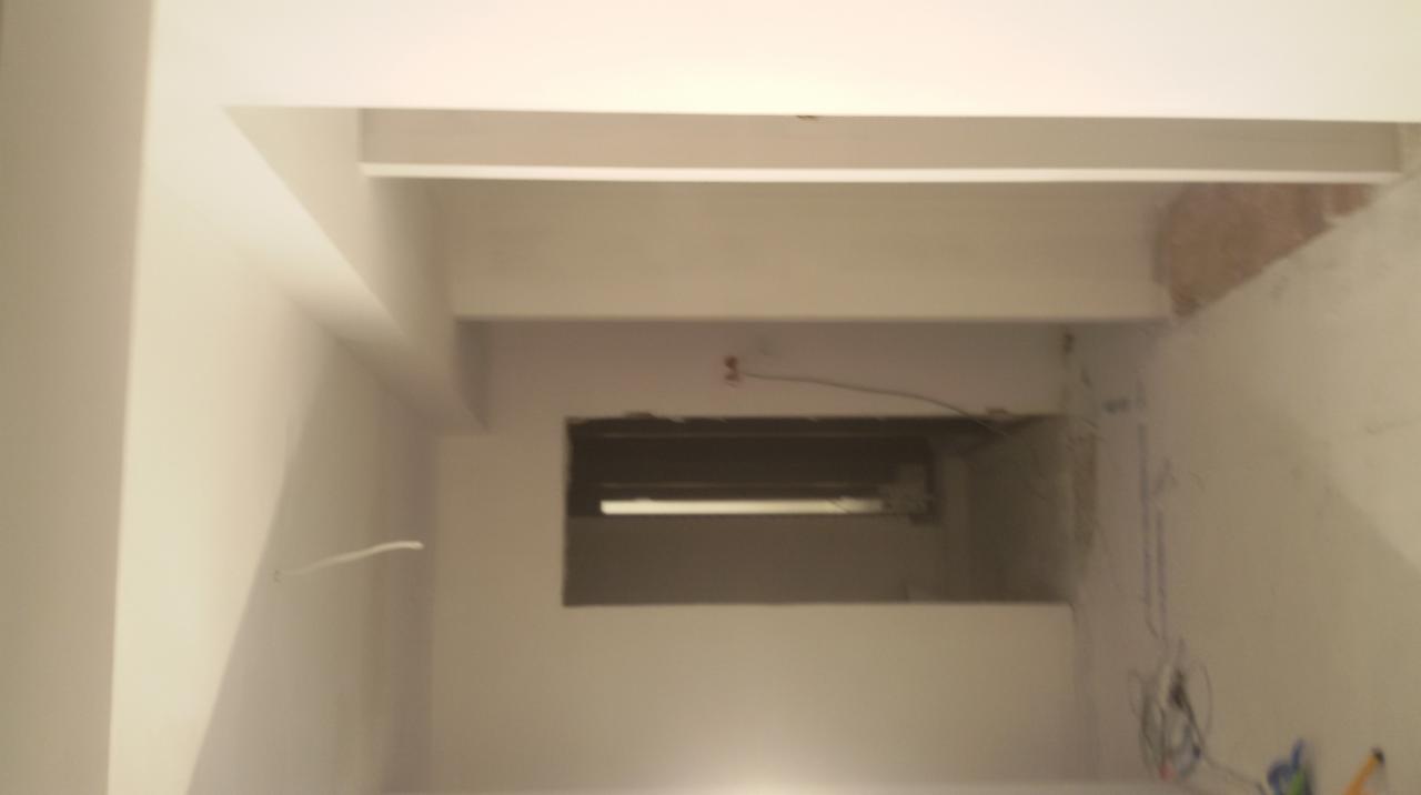 Wnętrze w trakcie prac wykończeniowych: gładkie, białe ściany z widocznymi przewodami i otworem w ścianie. Fragment surowej ściany w rogu. Oświetlenie sufitowe.