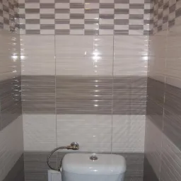 Wykończona toaleta z szarymi i białymi płytkami ułożonymi w poziome pasy i szachownicę na górze, widoczny biały sedes z metalową rurką doprowadzającą wodę.