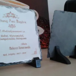 Dwa ozdobne, kamienne podziękowania na plastikowych podstawkach, jedno z dedykacją 'Kochanej Pani Magdzie', drugie puste, na tle ekspresu do kawy i ziarnistej grafiki.