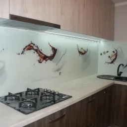 Szklany panel kuchenny z nadrukiem kawy i filiżanek, zamontowany nad blatem i kuchenką gazową w nowoczesnej kuchni z drewnianymi szafkami.