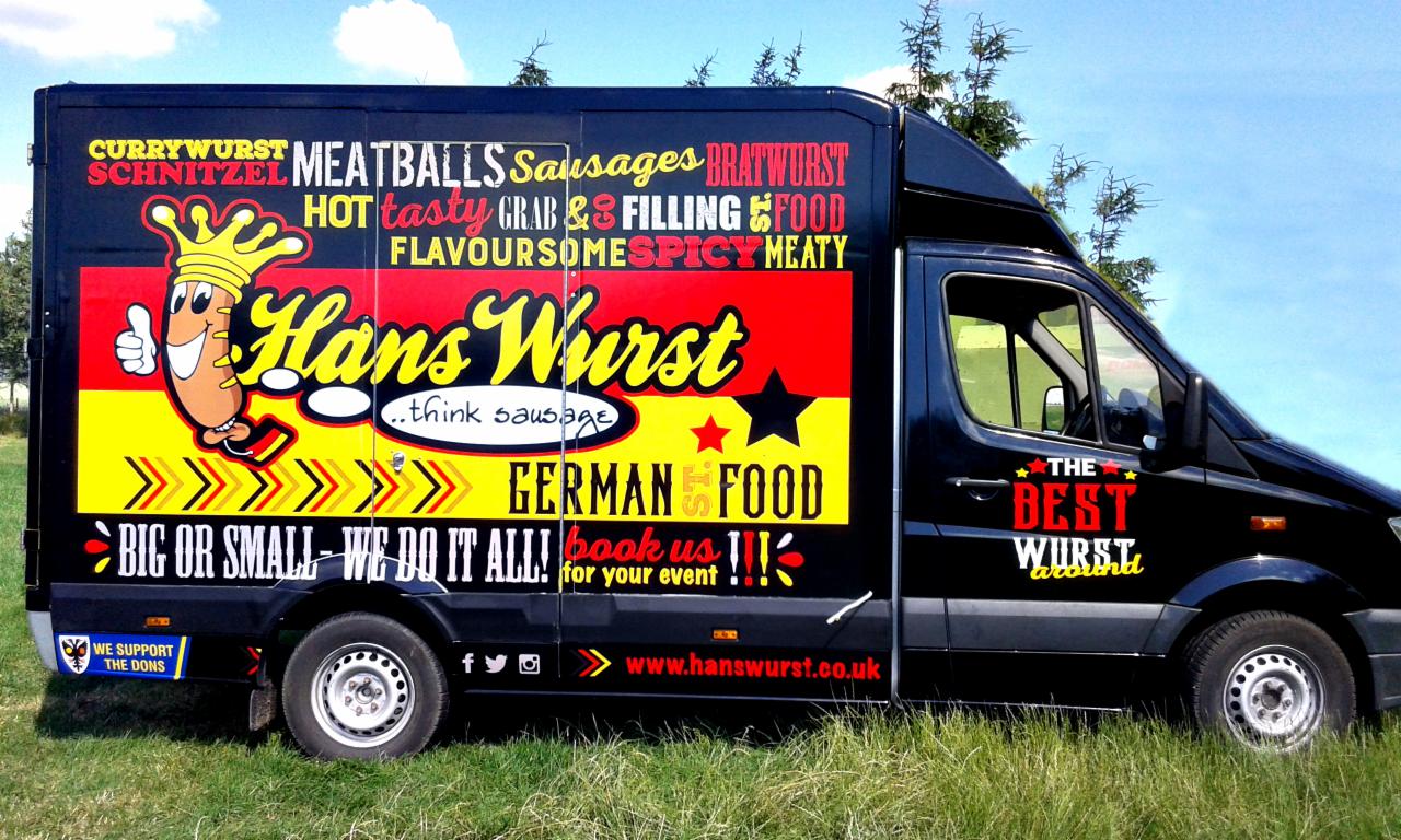 Czarny van z kolorową grafiką reklamową niemieckiego jedzenia, w tym wizerunkiem uśmiechniętej kiełbaski w koronie i napisami 'Hans Wurst', 'German Food', 'The Best Wurst around'.