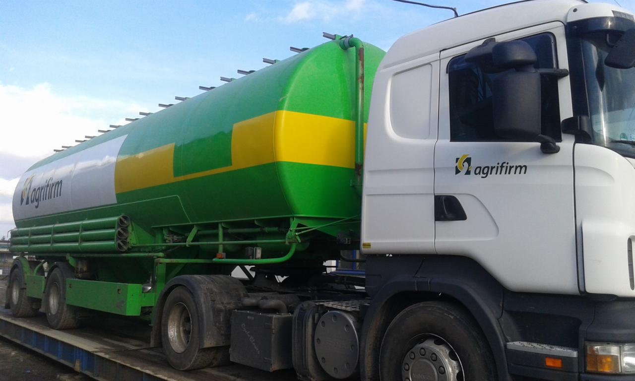 Ciężarówka z zielono-żółtym zbiornikiem, oznaczona logo 'Agrifirm', na tle pochmurnego nieba.