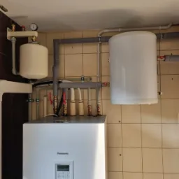 Biała pompa ciepła Panasonic w kotłowni z brązowo-beżowymi płytkami, widoczne rury i zbiornik wyrównawczy.