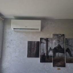 SM SYSTEM - Biała klimatyzacja Gree na ścianie z dekoracyjnym tynkiem, pod nią zestaw obrazów w szarościach z motywem futurystycznym. Widok z okna z roletami w lewym górnym rogu.