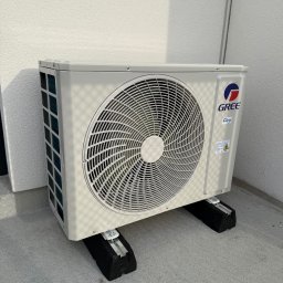 SM SYSTEM - Biała jednostka zewnętrzna klimatyzacji GREE na czarnych podkładkach antywibracyjnych, zamontowana przy jasnej ścianie budynku. Widoczne detale montażu.