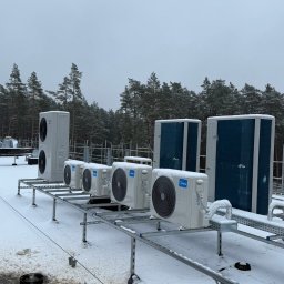 SM SYSTEM - Białe jednostki zewnętrzne klimatyzacji Midea na metalowej konstrukcji, pokryte śniegiem, na dachu budynku. W tle las iglasty i pochmurne niebo.