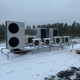 SM SYSTEM - Rząd jednostek zewnętrznych klimatyzacji Midea EasyFit na dachu pokrytym śniegiem, z lasem w tle. Widoczne ślady stóp w śniegu.