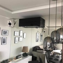 SM SYSTEM - Czarny klimatyzator ścienny w nowoczesnym salonie z galerią czarno-białych zdjęć, lampami wiszącymi i lustrem. Widoczne głośniki na ścianie.