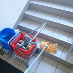 Kamil Rybiński- Usługi GREENI - Wózek ze sprzętem do sprzątania (mop, wiadra) na tle schodów z płytek. Mop leży na schodku, gotowy do użycia. Widoczne ślady użycia sprzętu.