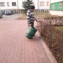 Kamil Rybiński- Usługi GREENI - Mężczyzna w czapce i bluzie w paski zbiera liście do zielonego worka przy krzewach na tle bloku mieszkalnego.