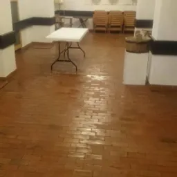 Sala z ceglaną podłogą po umyciu, widoczne białe stoły i krzesła w tle, oświetlona żyrandolem.