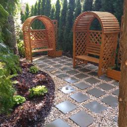 Kamil Rybiński- Usługi GREENI - Dwa drewniane łuki z ławkami, ustawione w symetrycznym ogrodzie z kamienną ścieżką i rabatami z roślinami ozdobnymi, na tle wysokich, zielonych drzew.