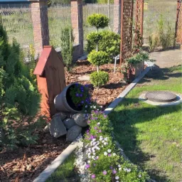 Aranżacja ogrodu z przewróconą donicą, kamieniami i rabatą kwiatową w odcieniach fioletu i bieli, na tle ogrodzenia z ceglanymi słupkami i drewnianej pergoli.