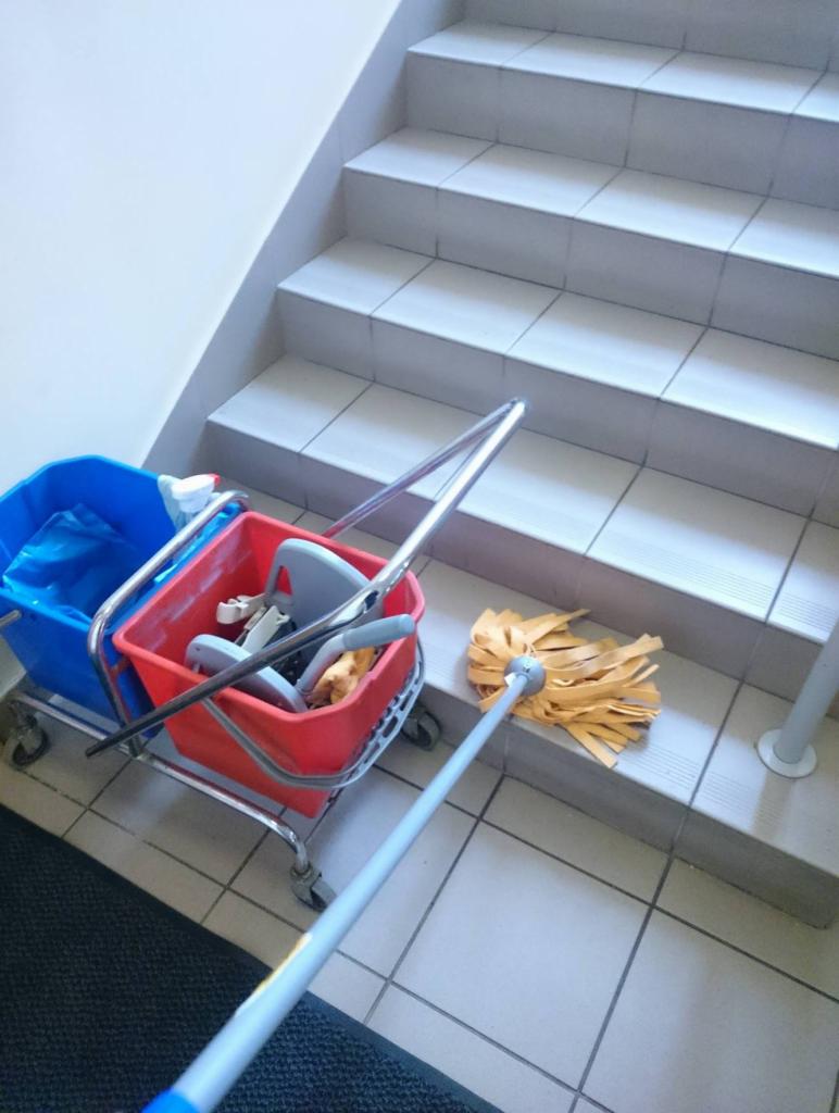 Wózek ze sprzętem do sprzątania (mop, wiadra) na tle schodów z płytek. Mop leży na schodku, gotowy do użycia. Widoczne ślady użycia sprzętu.