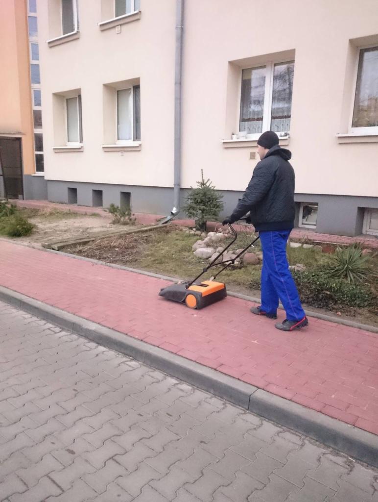 Mężczyzna w niebieskich spodniach zamiata chodnik przed budynkiem mieszkalnym za pomocą pomarańczowo-czarnej zamiatarki mechanicznej.