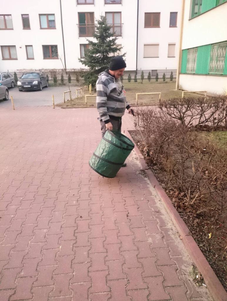 Mężczyzna w czapce i bluzie w paski zbiera liście do zielonego worka przy krzewach na tle bloku mieszkalnego.