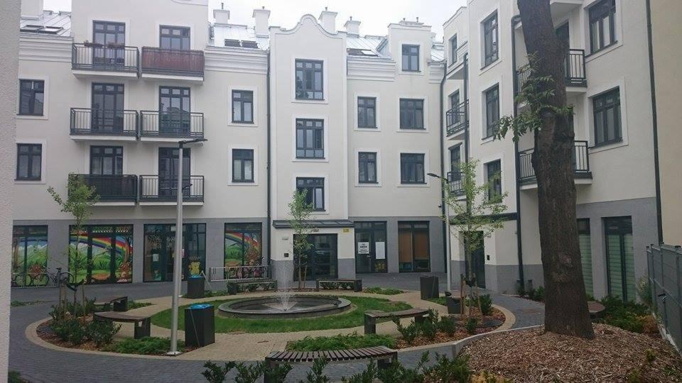 Dziedziniec nowoczesnego apartamentowca z fontanną otoczoną zielenią, ławkami i młodymi drzewkami; w tle budynek w jasnej kolorystyce z balkonami i lokalami usługowymi na parterze.