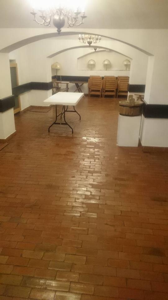 Sala z ceglaną podłogą po umyciu, widoczne białe stoły i krzesła w tle, oświetlona żyrandolem.
