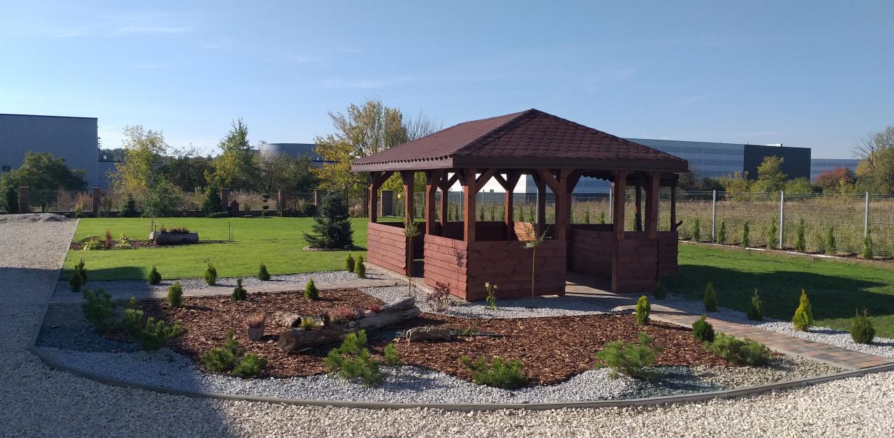 Drewniana altana z brązowym dachem w zadbanym ogrodzie z ozdobnymi nasadzeniami i korą, na tle zielonego trawnika i budynków przemysłowych w oddali.