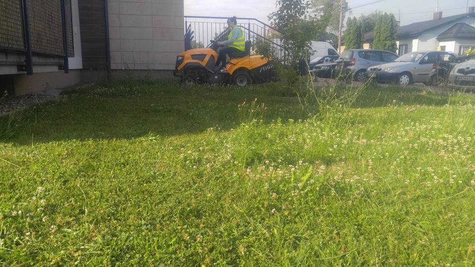 Żółta kosiarka traktorek Stiga w trakcie koszenia zarośniętego trawnika przed budynkiem z metalową barierką.