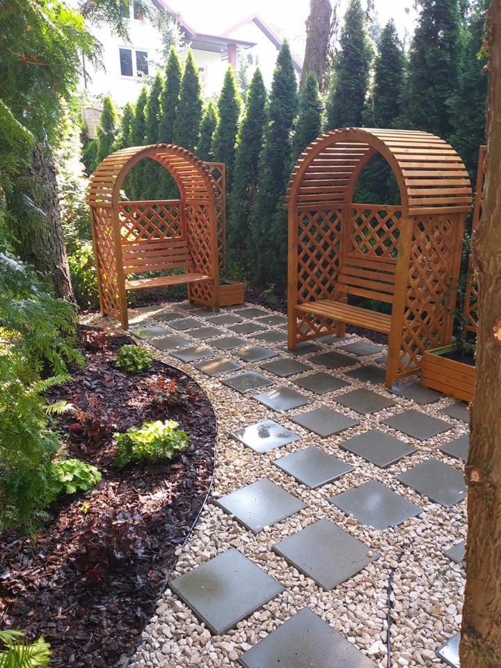 Dwa drewniane łuki z ławkami, ustawione w symetrycznym ogrodzie z kamienną ścieżką i rabatami z roślinami ozdobnymi, na tle wysokich, zielonych drzew.