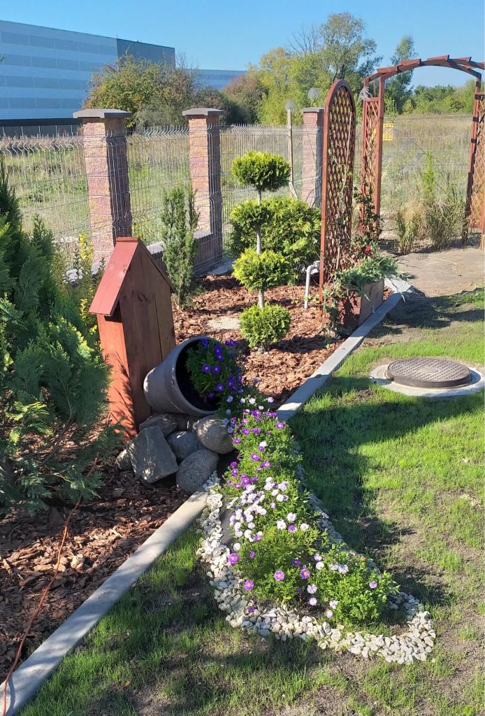 Aranżacja ogrodu z przewróconą donicą, kamieniami i rabatą kwiatową w odcieniach fioletu i bieli, na tle ogrodzenia z ceglanymi słupkami i drewnianej pergoli.