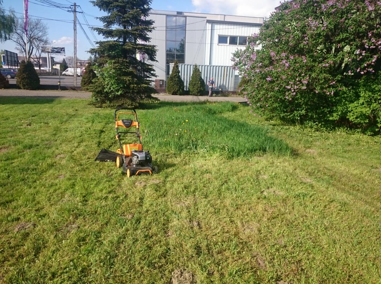 Pomarańczowo-czarna kosiarka spalinowa na trawniku z częściowo skoszoną trawą, w tle budynek biurowy i osoba przy kontenerze.
