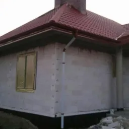 Dom w trakcie budowy z czerwoną dachówką i oknami zabezpieczonymi folią. Widoczne białe bloczki i rynna spustowa. Etap surowy zamknięty.