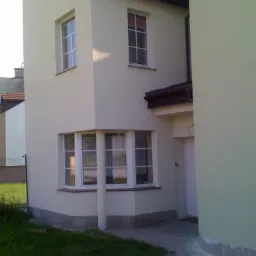 Jasna elewacja domu jednorodzinnego z oknami z białymi ramami. Widoczny fragment dachu i trawnik. Ujęcie z poziomu gruntu, podkreślające bryłę budynku.